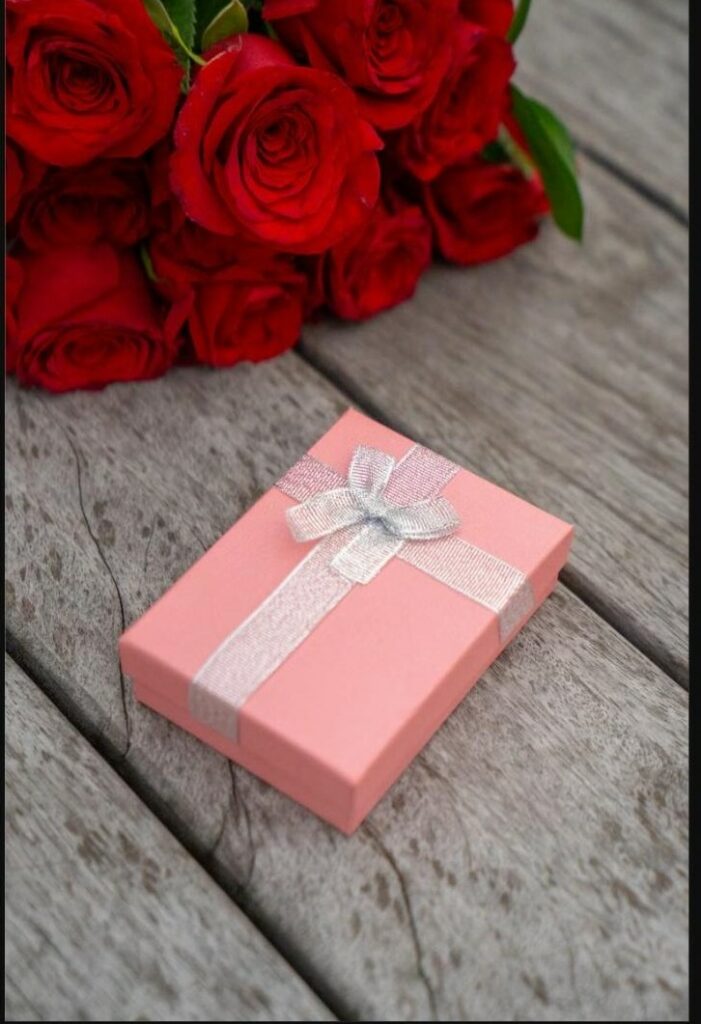 Petite boîte cadeau rose avec ruban près d'un bouquet de roses rouges, symbole d'une surprise à découvrir en couple